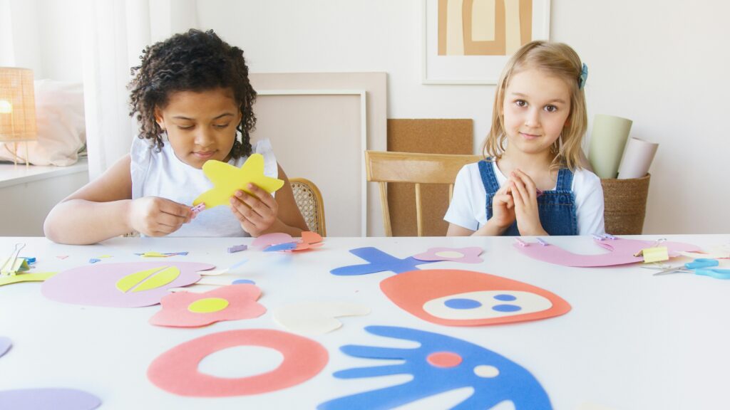 Two children happily participate in an arts and crafts activity, creating colorful shapes indoors.